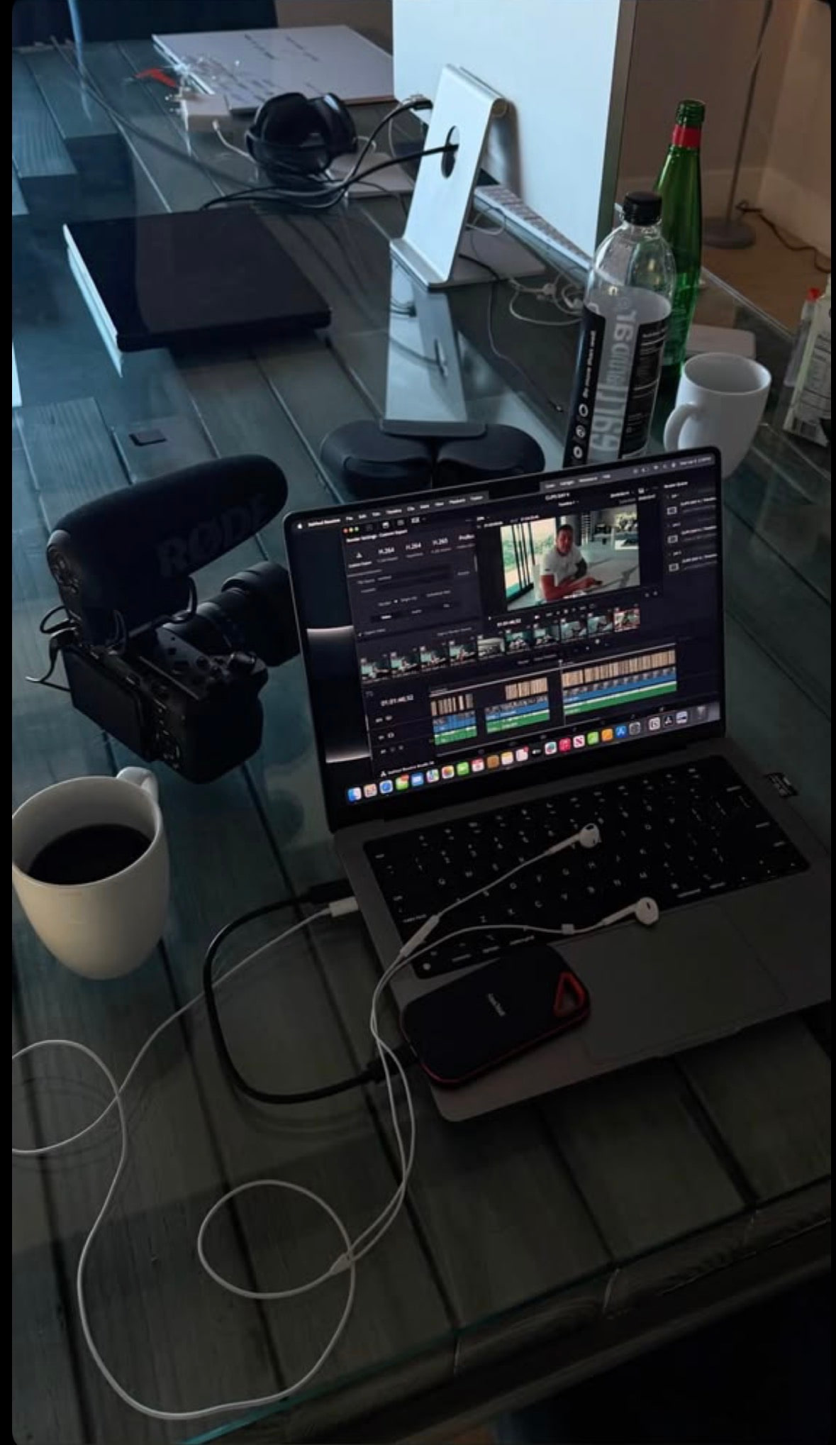 A laptop running a video edit at a glass desk, with a camera, microphone and a cup of coffee.