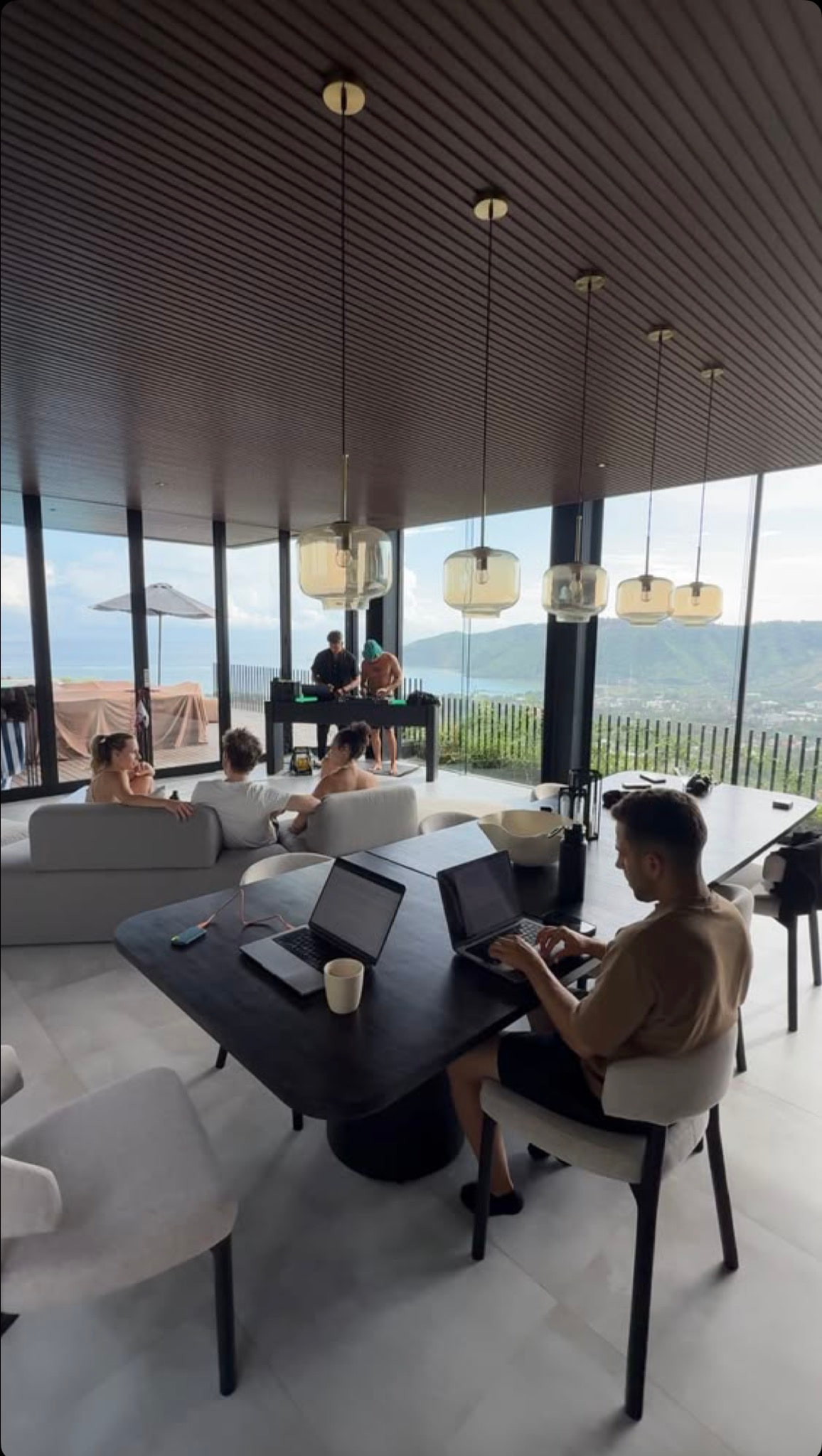 People working on laptops and talking in a bright open living space with floor-to-ceiling windows over the sea.