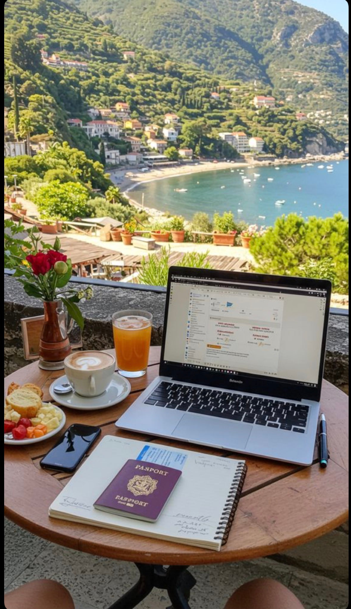 A laptop, notebook, passport and coffee on an outdoor table overlooking a coastal town and bay.